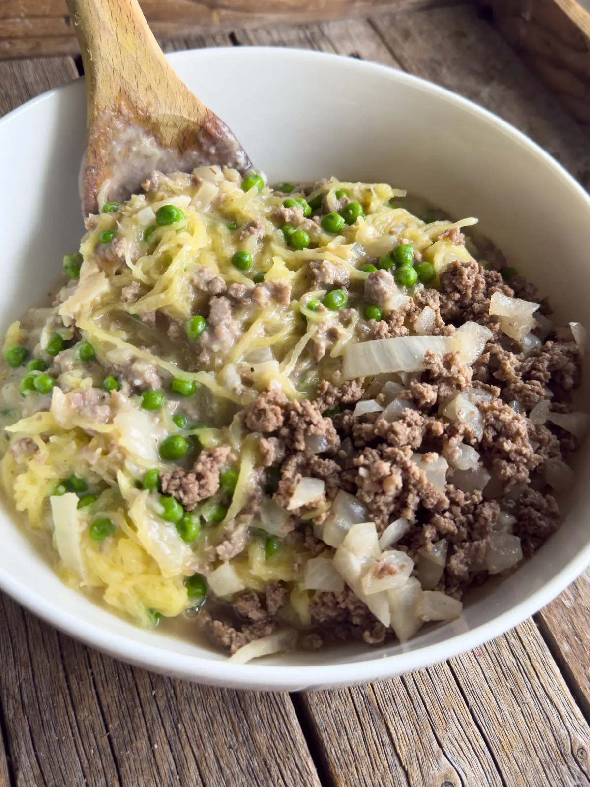 Creamy garlic spaghetti squash with ground beef and peas in a large bowl with a wooden spoon in it.