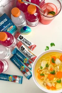 A birds-eye view of a bowl of chicken soup on a counter next to electrolyte drinks and packets.