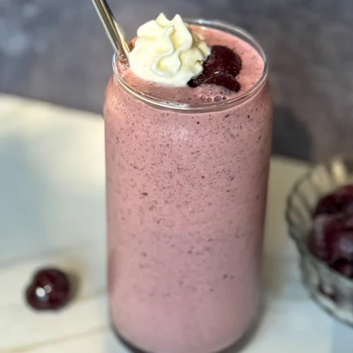 A dark cherry cottage cheese smoothie in a tall glass with a straw in it garnished with dark cherries and whipped cream on a counter next to a bowl of cherries.