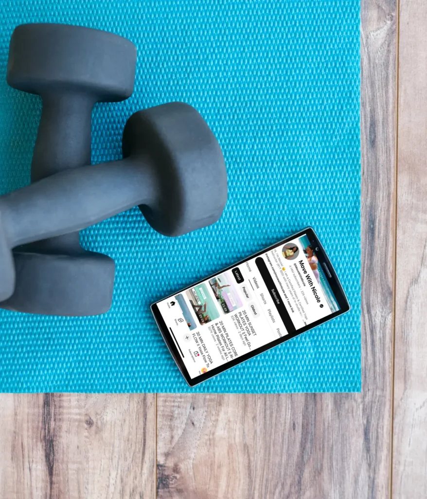 A birds-eye view of a set of dumbbells on a yoga mat next to a cell phone with YouTube pulled up.