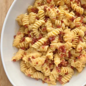 A birds-eye view of the butternut squash pasta sauce with rotini noodles and crispy pancetta in a white bowl on a cutting board.