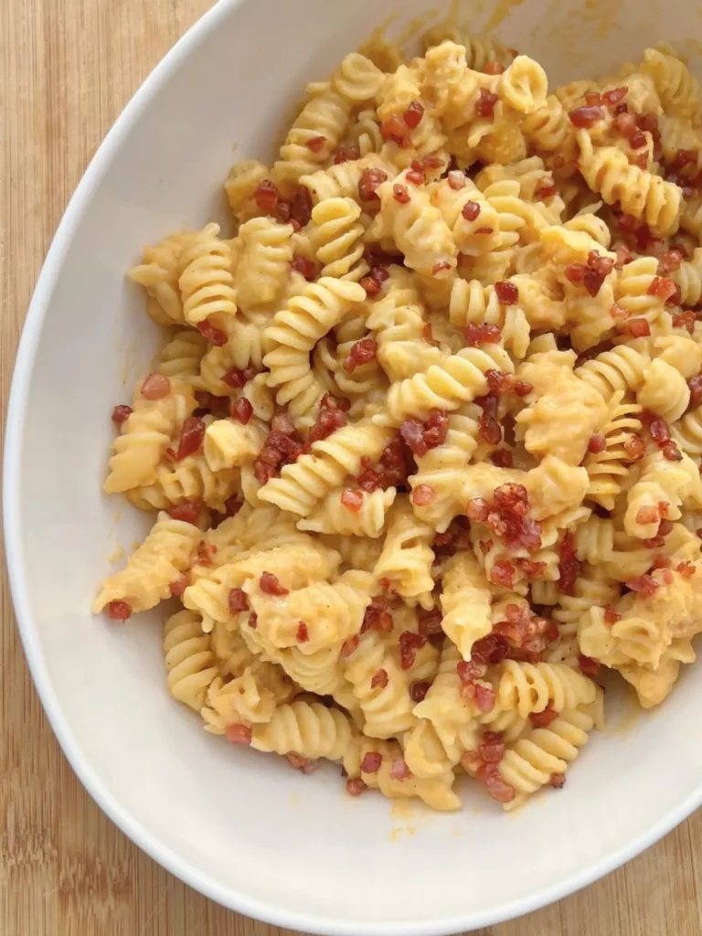 A birds-eye view of the butternut squash pasta sauce with rotini noodles and crispy pancetta in a white bowl on a cutting board.