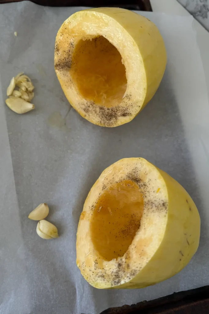 Two spaghetti squash halves next to two garlic cloves on a baking sheet lined with parchment paper.