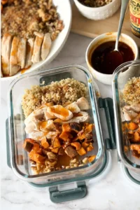Two meal prep storage containers with sweet potatoes, chicken and quinoa on a counter next to meal prep ingredients.