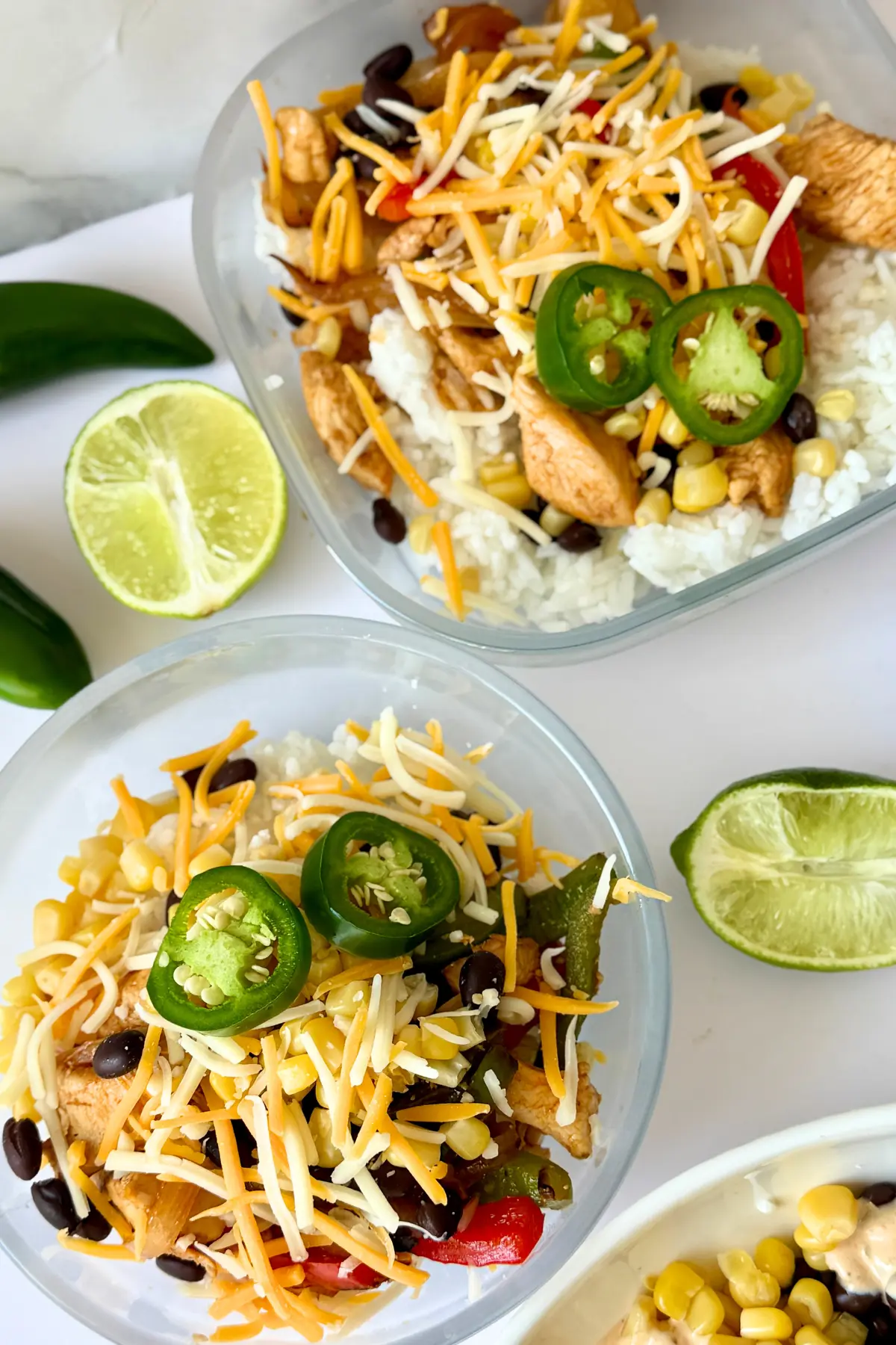 Two meal prep storage containers with chicken, veggies, balck beans, corn, shredded cheese and rice on a counter.