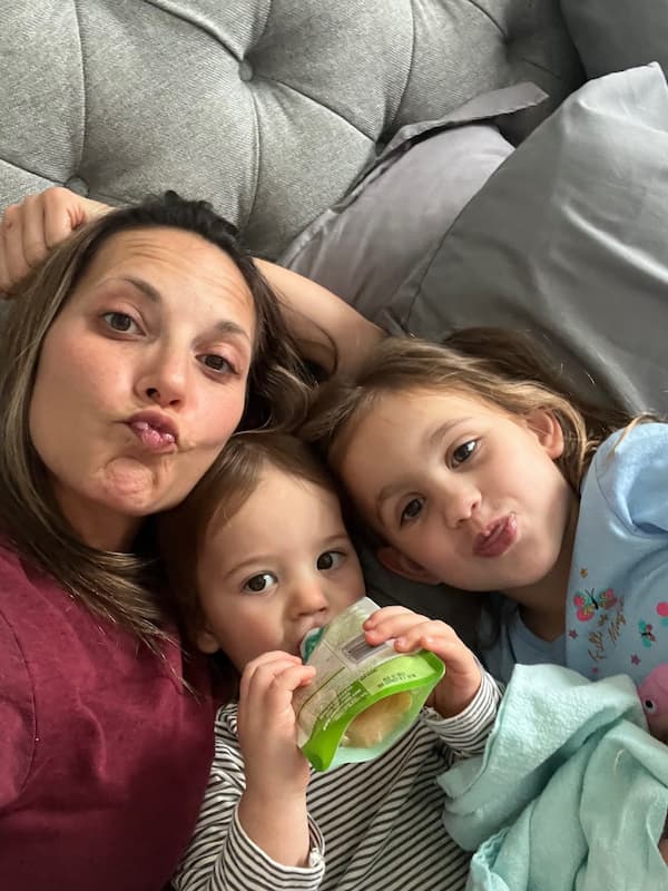 A stay-at-home mom in bed with her two girls making funny faces.