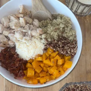 A fully assembled quinoa bowl with butternut squash cubes, chopped pecans, parmesan cheese, diced chicken and bacon in a large bowl on a wooden cutting board.