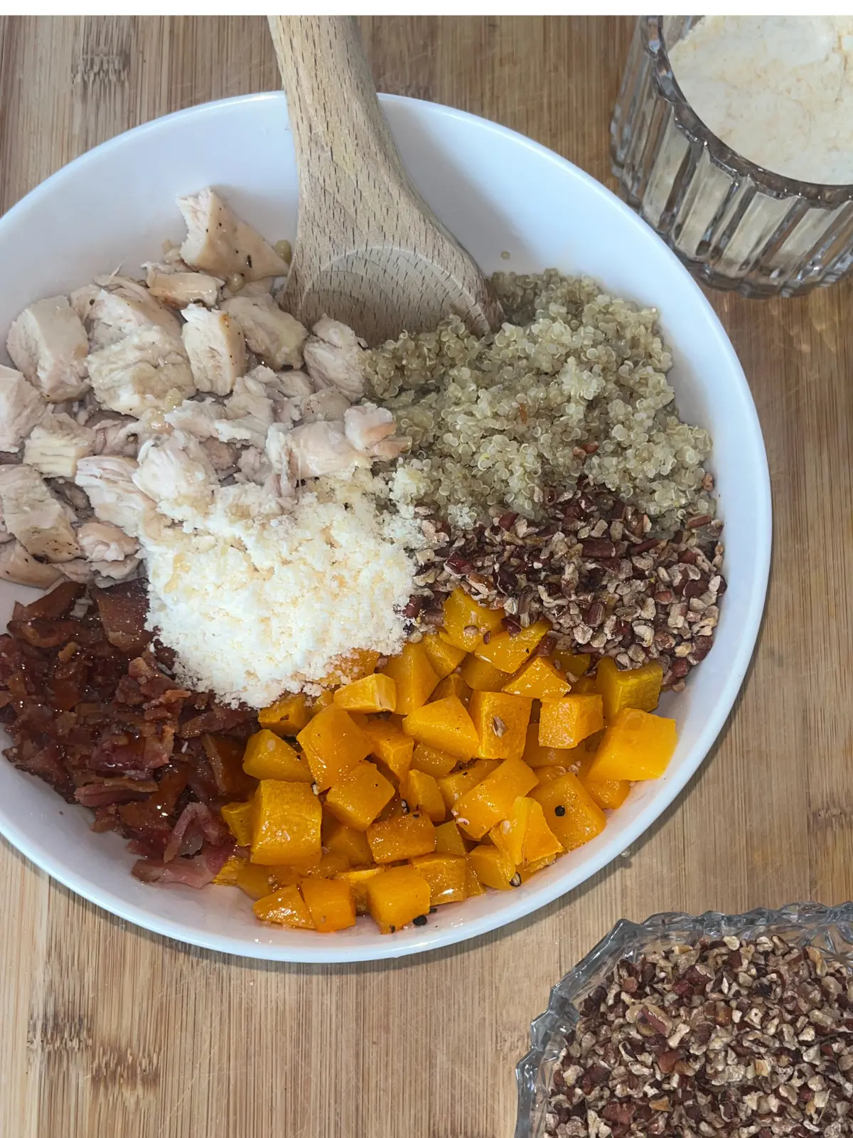 A fully assembled quinoa bowl with butternut squash cubes, chopped pecans, parmesan cheese, diced chicken and bacon in a large bowl on a wooden cutting board.