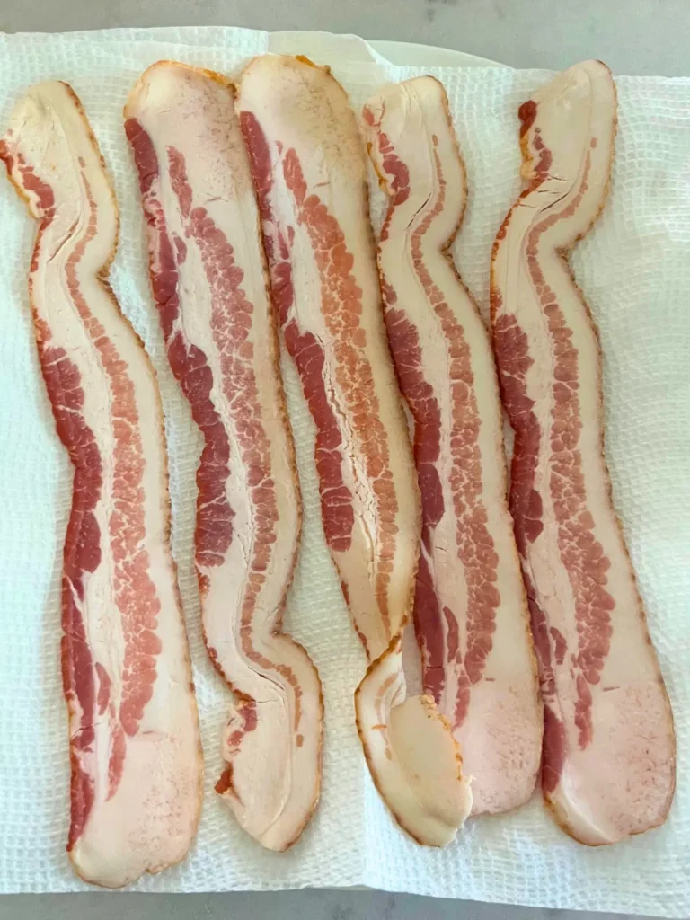 5 slices of raw bacon on a plate lined with paper towel.