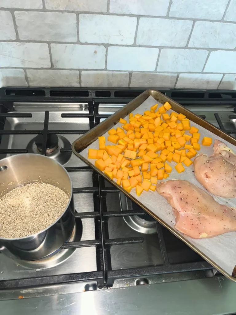 A pot of quinoa cooking on the stove next to a baking sheet lined with parchment paper with butternut squash cubes and raw chicken breast.