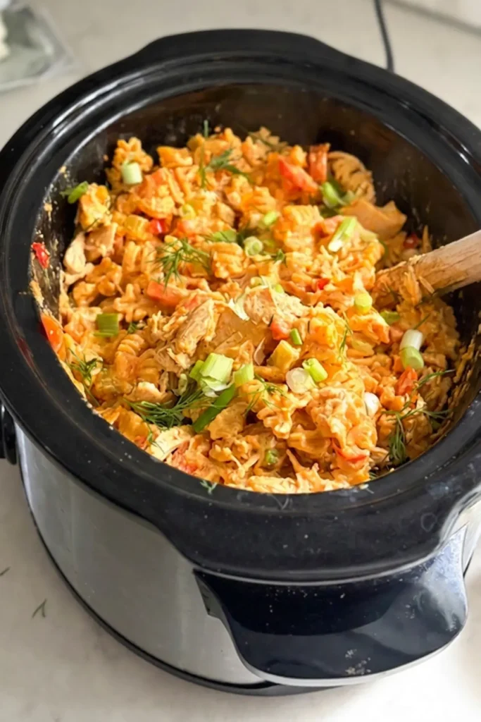 A birds-eye view of the cooked buffalo chicken pasta in a slow cooker with a wooden spoon in it, garnished with green onions.