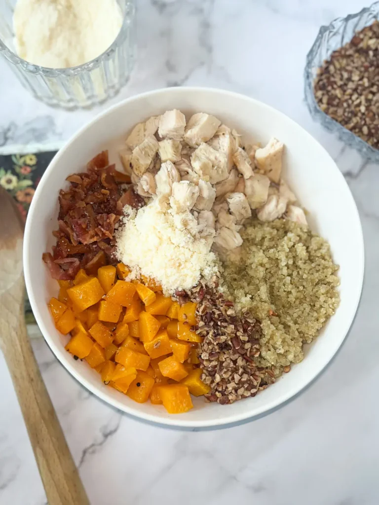 A large bowl with roasted butternut squash cubes, diced chicken, bacon, chopped pecans, quinoa and parmesan cheese on a counter next to a wooden spoon.