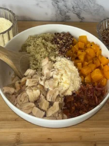 Roasted butternut squash, quinoa, parmesan cheese, bacon, and diced chicken in a large white bowl drizzled with honey with a wooden spoon in it.