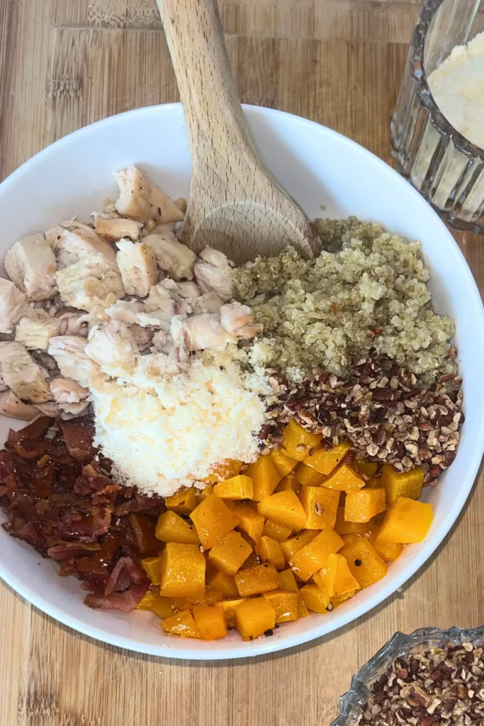 A birds-eye view of quinoa, butternut squash, bacon, parmesan and pecans in a bowl with a wooden spoon in it.
