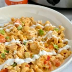A big bowl of buffalo chicken pasta drizzled with ranch dressing next to a bottle of buffalo sauce and a crockpot.