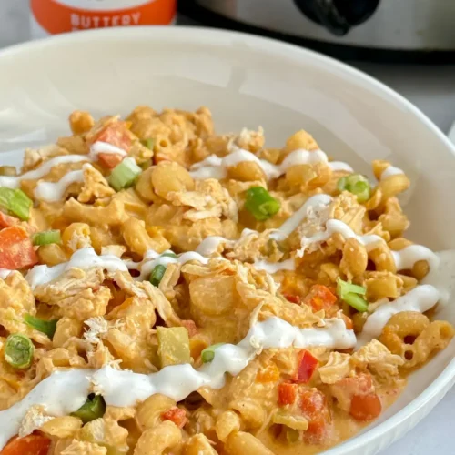 A big bowl of buffalo chicken pasta drizzled with ranch dressing next to a bottle of buffalo sauce and a crockpot.
