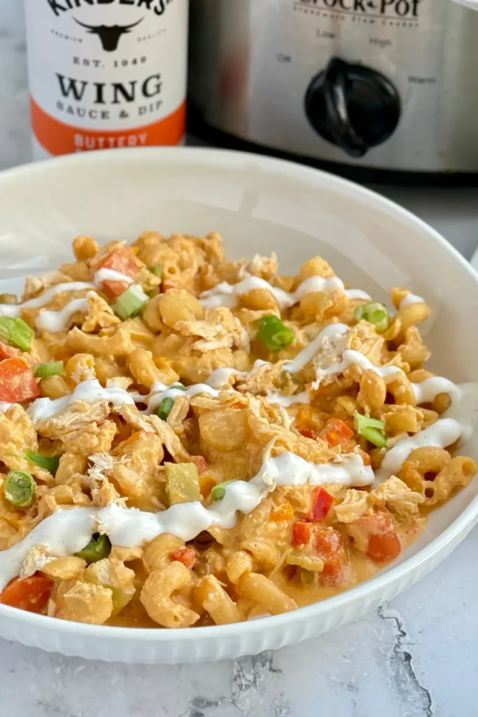 A big bowl of buffalo chicken pasta drizzled with ranch dressing next to a bottle of buffalo sauce and a crockpot.