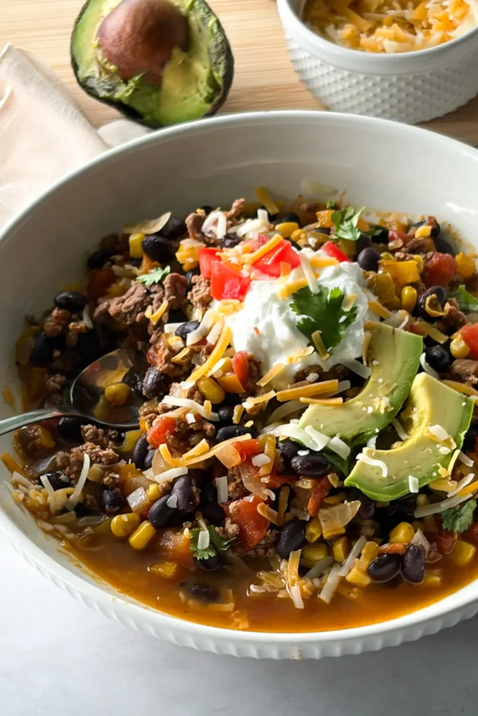 A close-up of a bowl of Mexican chili topped with yogurt and avocado slices next to an avocado half.