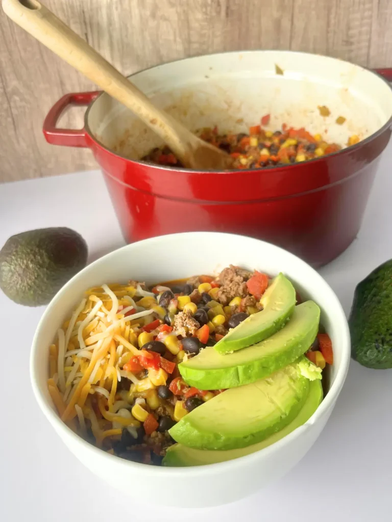 A bowl of Mexican chili next to a pot of chili on a counter next to avocados. 