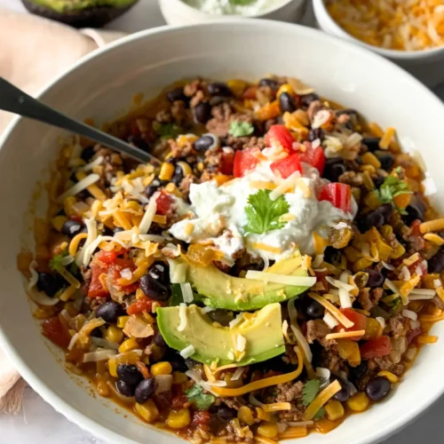 A close-up of a bowl of Mexican chili topped with yogurt and avocado slices next to an avocado half.