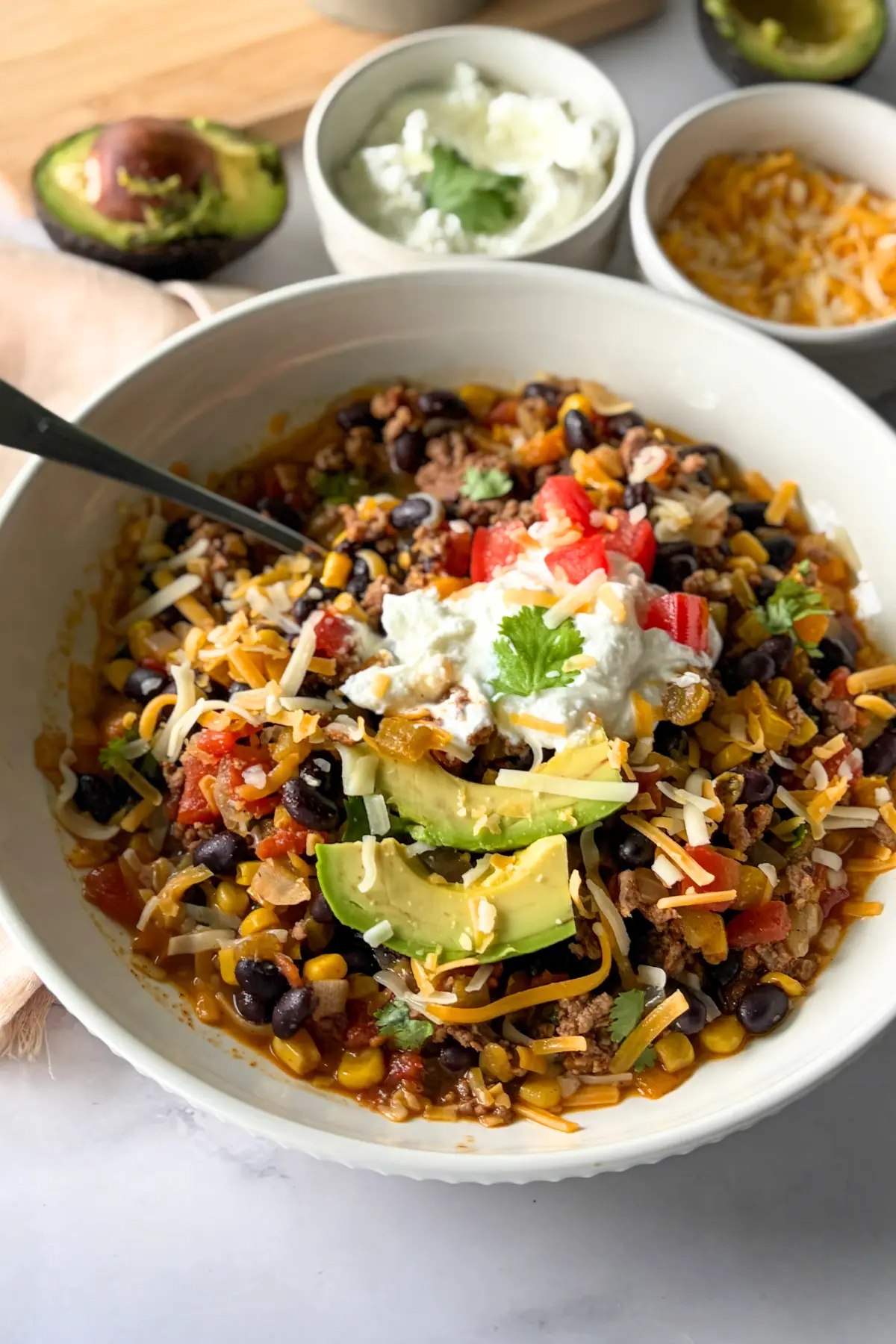 A close-up of a bowl of Mexican chili topped with yogurt and avocado slices next to an avocado half.