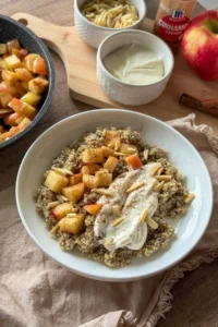 An apple quinoa breakfast bowl on a table next to apples, yogurt, and a cutting board.
