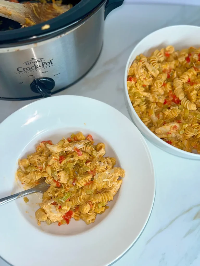 High protein buffalo chicken pasta with cottage cheese in a bowl with a fork next to a crockpot and large bowl of pasta on the counter.