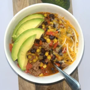 A bowl of Mexican-inspired chili topped with cheese and avocado slices on a wooden board next to an avocado.