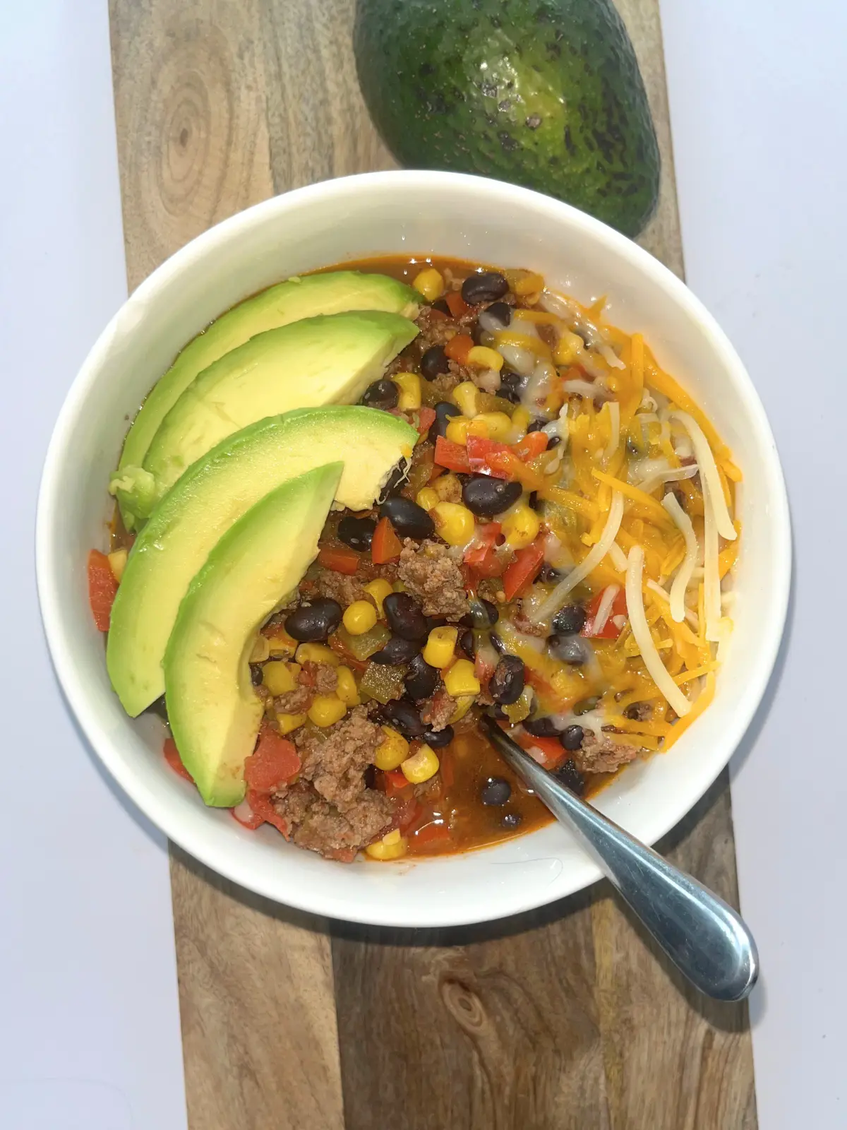 A bowl of Mexican-inspired chili topped with cheese and avocado slices on a wooden board next to an avocado.