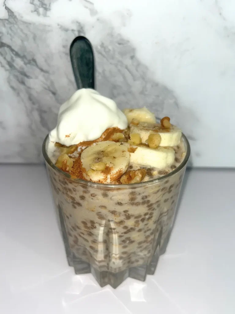 High protein banana cinnamon overnight oats in a glass cup with a spoon in it, topped with whipped cream and banana slices.