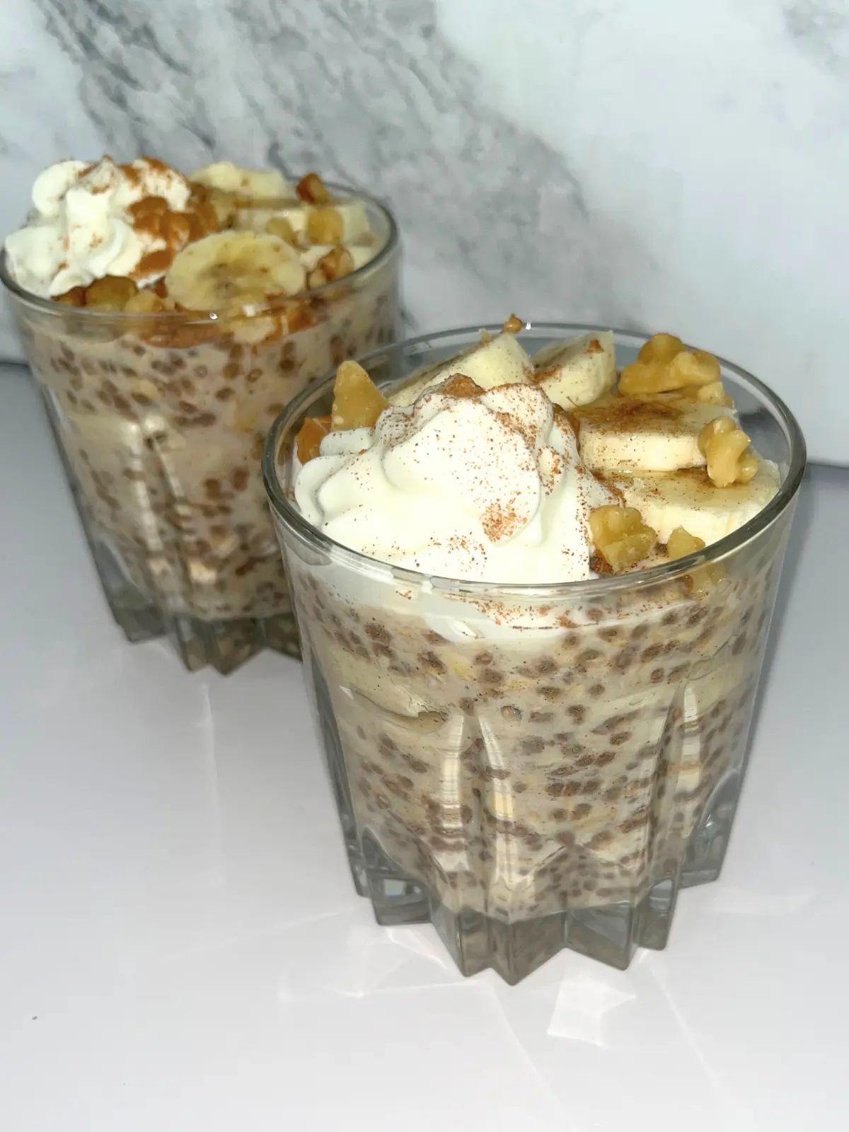 Banana cinnamon overnight oats with protein powder in two glass cups topped with whipped cream, banana slices and chopped nuts.