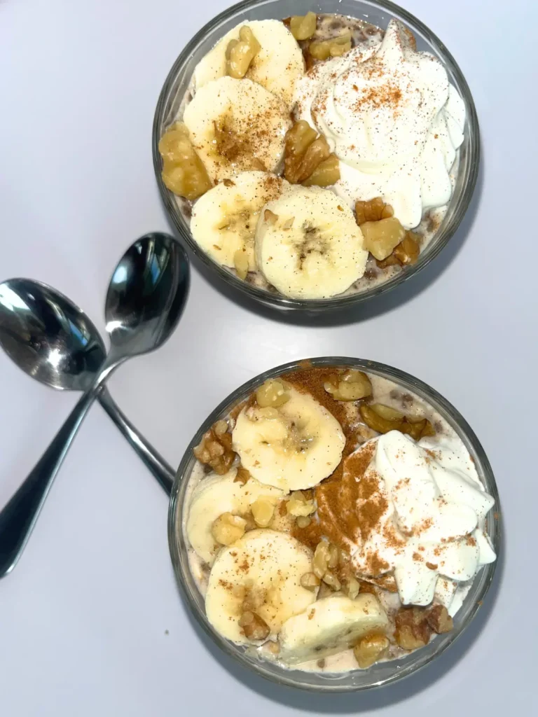 A birds-eye view of two high protein banana cinnamon overnight oats topped with extra cinnamon, whipped cream and banana slices on a counter next to spoons.