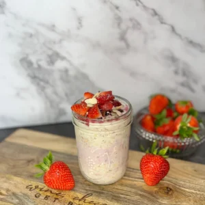 Overnight oats topped with strawberries and blueberries on a wood cutting board on the counter next to a bowl of strawberries.