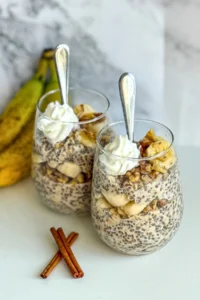 Two glasses of banana cinnamon overnight oats topped with whipped cream and walnuts on a counter next to cinnamon sticks and bananas.