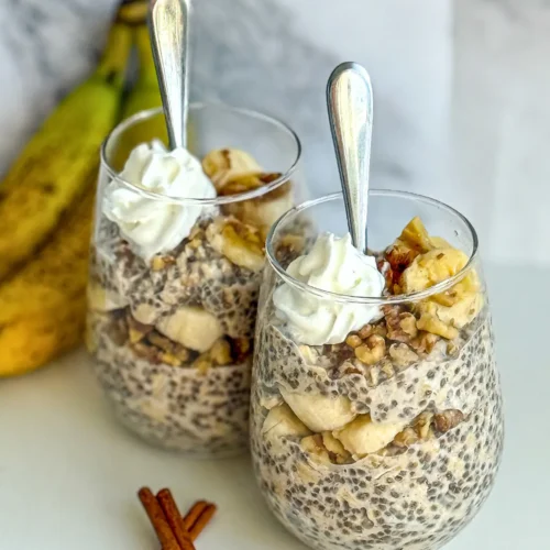 Two glasses of banana cinnamon overnight oats topped with whipped cream and walnuts on a counter next to cinnamon sticks and bananas.