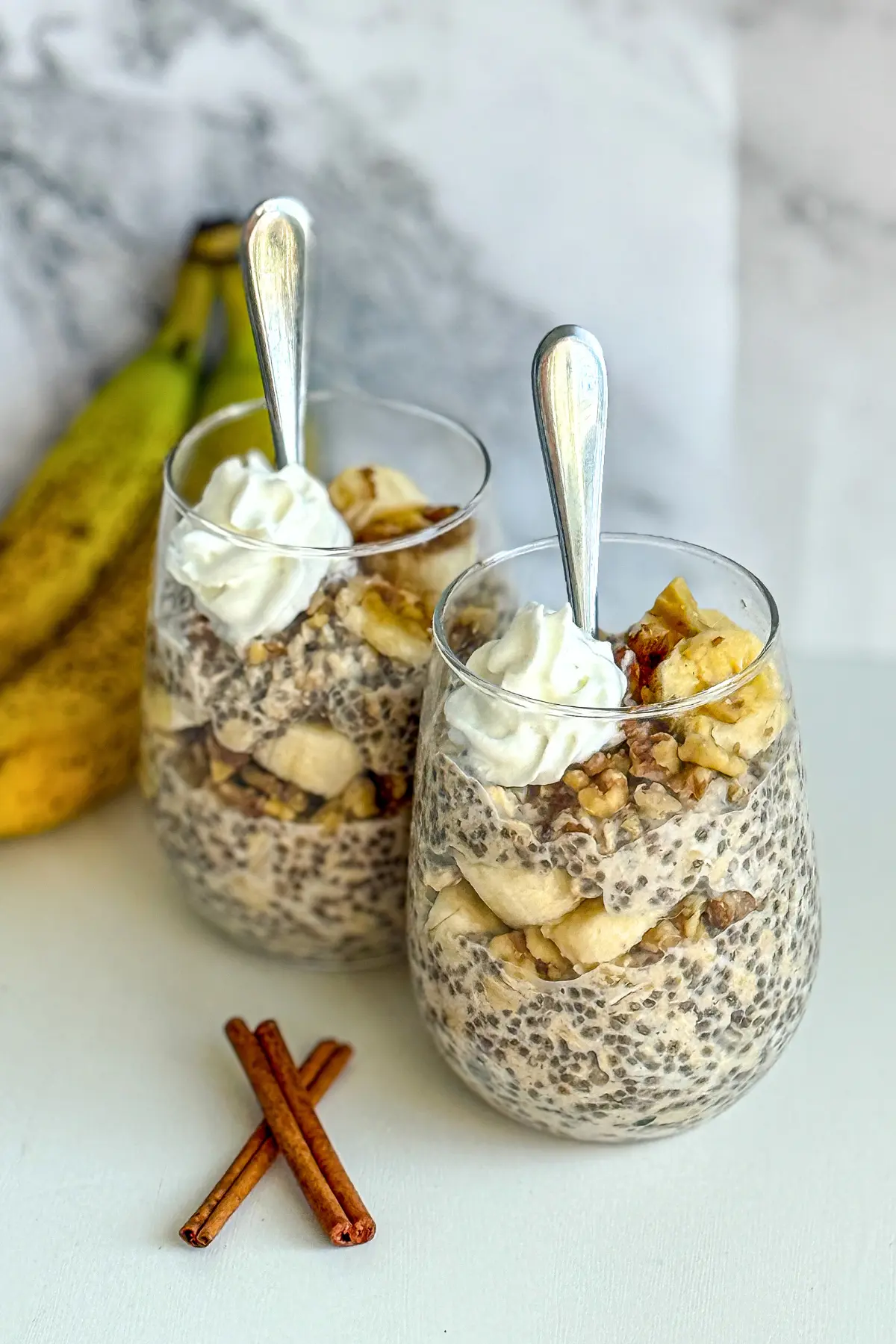 Two glasses of banana cinnamon overnight oats topped with whipped cream and walnuts on a counter next to cinnamon sticks and bananas.