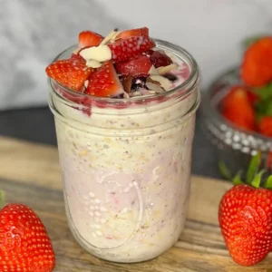 Overnight oats topped with strawberries and blueberries on a wood cutting board on the counter next to a bowl of strawberries.