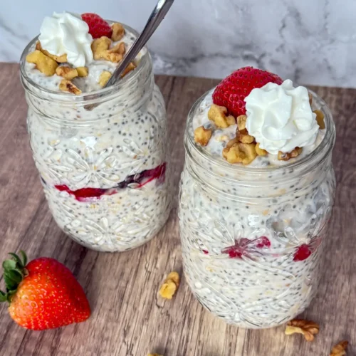 Berry cheesecake overnight oats with protein powder in two large mason jars topped with whipped cream and chopped nuts on a counter.