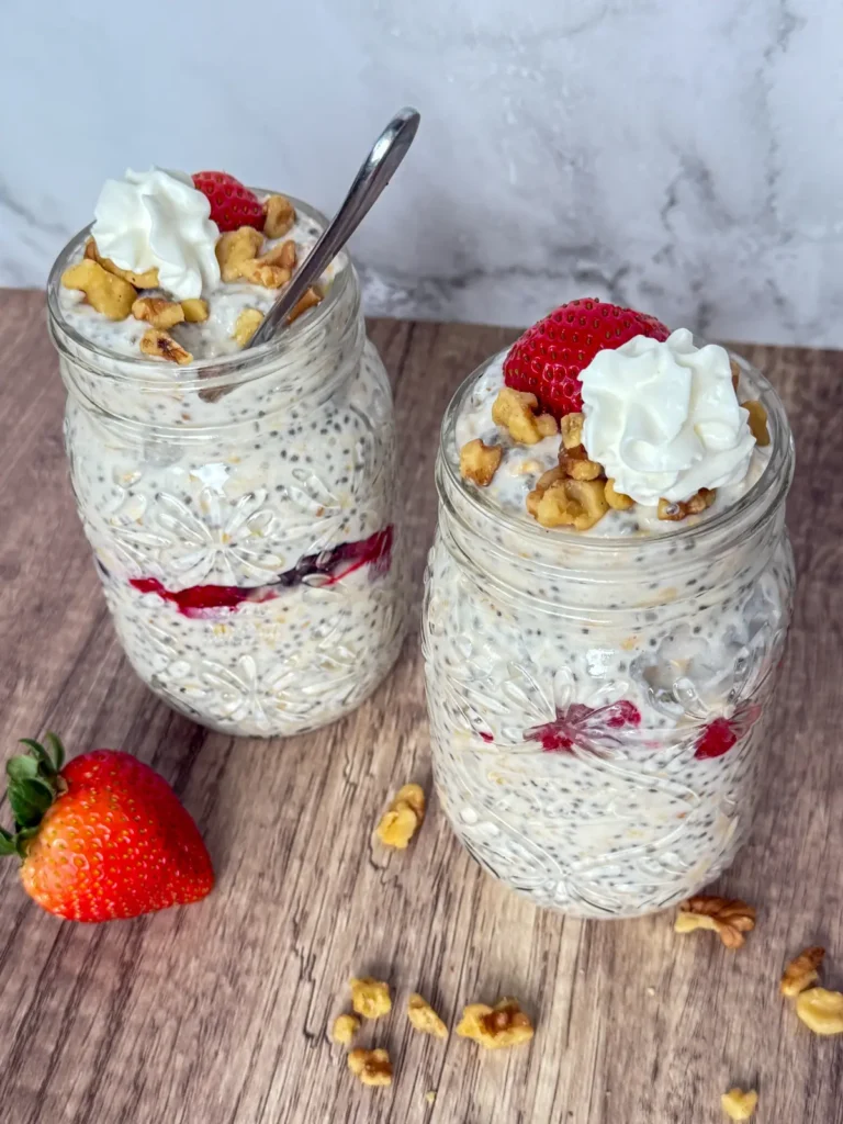 Berry cheesecake overnight oats with protein powder in two large mason jars topped with whipped cream and chopped nuts on a counter.