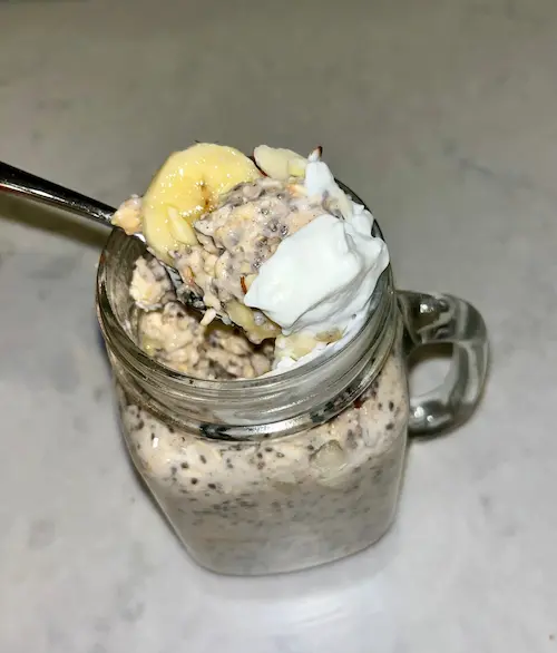 A masor jar of banana cinnamon overnight oats topped with whipped cream with a spoon in it.