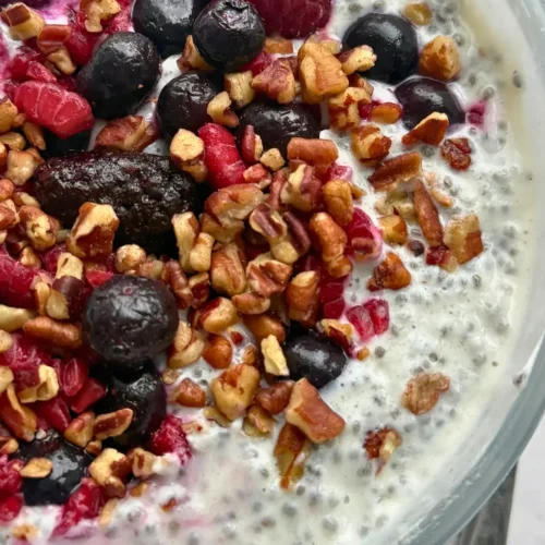A chia breakfast bowl topped with chopped pecans, fruit and honey on a counter next to a spoon.