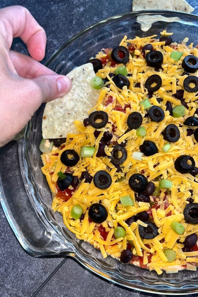 A fully assembled 7-layer high protein taco dip topped with cheese, green onions, and olives with someone dipping a tortilla chip into it. 