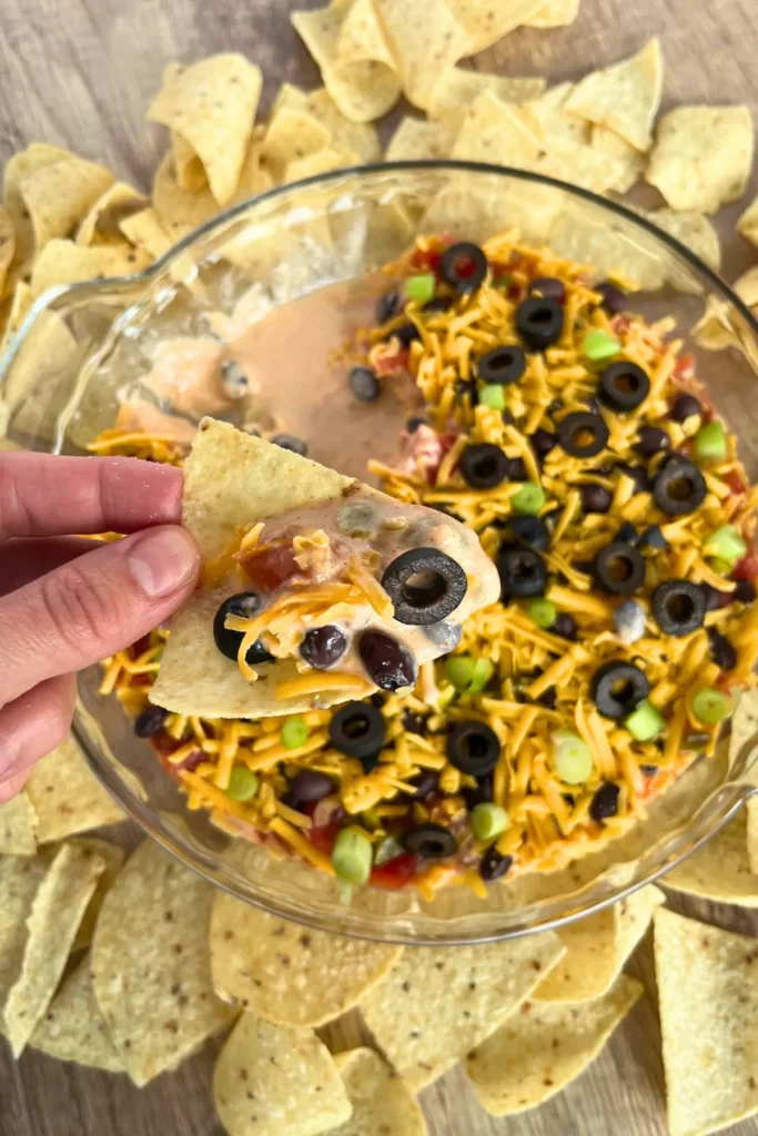 Someone dipping a chip into the 7-layer high protein taco dip which is on a cutting board surrounded by chips.
