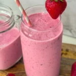 A creamy strawberry gut health smoothie with kefir in a tall glass with a pink straw in it, garnished with a strawberry on a wooden platter.