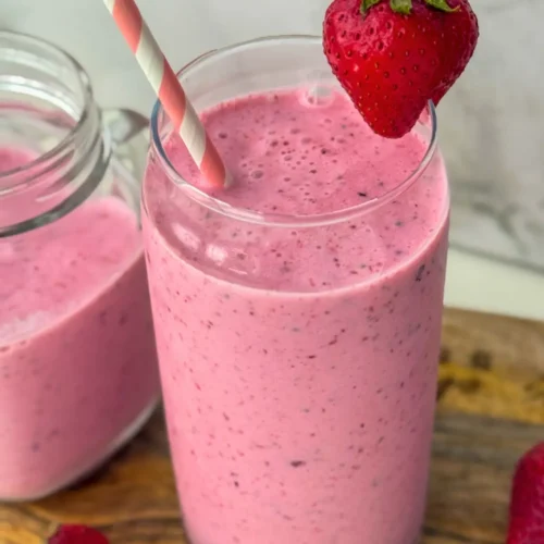 A creamy strawberry gut health smoothie with kefir in a tall glass with a pink straw in it, garnished with a strawberry on a wooden platter.