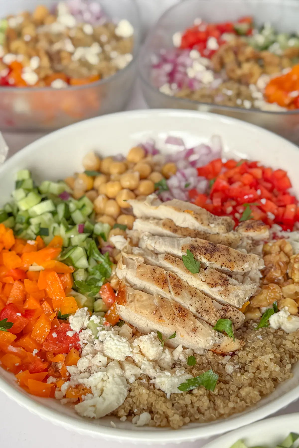 A colorful, fully assembled Mediterranean quinoa bowl with each vegetable and component separated on a counter with two meal prep bowls in the background.