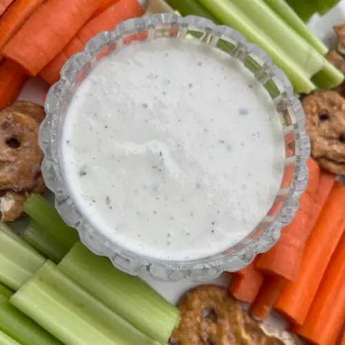 A glass dish of the health ranch dip surrounded by celery, carrots, and pretzels on a white platter.