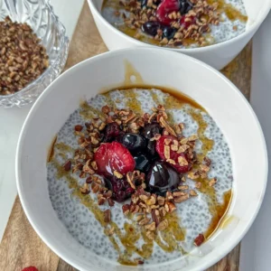 Two high protein and fiber chia breakfast bowls on a wooden cutting board, topped with fruit, chopped nuts and a drizzle of honey.