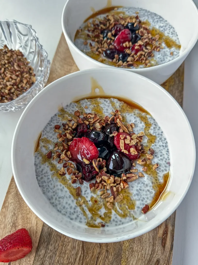 Two high protein and fiber chia breakfast bowls on a wooden cutting board, topped with fruit, chopped nuts and a drizzle of honey.