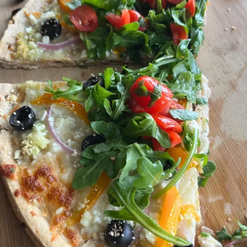 A close-up of two slices of Mediterranean diet pizza on a wooden cutting board after slicing.